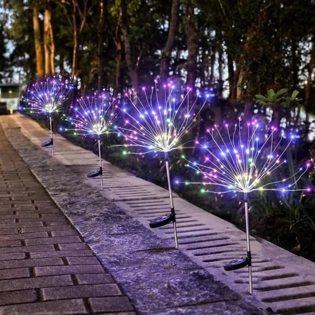 Two dandelion-style outdoor stake lights glowing in a grassy yard at night, with bright white “spark” branches and trees silhouetted in the background.