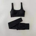 Black ribbed sports bra and matching high-waisted leggings set laid flat on a neutral gray background. The outfit is designed for activewear or athleisure, featuring a scoop neckline and wide straps on the bra.