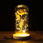 Glowing decorative lamp featuring a gold rose inside a clear glass dome, wrapped with warm white fairy lights on a wooden base against a dark background.