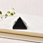 A minimalist close-up photo of a small glossy black pyramid-shaped crystal/stone placed on top of a light-colored fabric-covered book, with a soft neutral background and a few green leaves blurred in the corner.