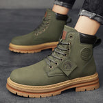 A pair of sleek black leather ankle boots with rugged rubber soles, lace-up fronts, and metal eyelets, displayed on a gray fabric surface with one boot propped up on a vintage-style book for presentation.