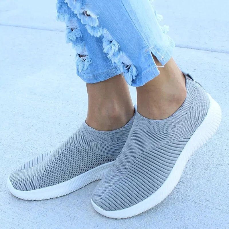Light grey slip-on knit trainers with white soles, worn with distressed light blue jeans.