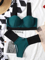 Teal bikini set laid flat on a bed, featuring a structured underwire top with wide black straps and a matching high-cut bottom with a wide black waistband, surrounded by magazines and fabric.