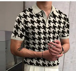 Person shown from shoulders to waist wearing a short-sleeve black-and-white houndstooth knit polo with a light collar and sleeve cuffs, standing indoors with hands clasped and a small forearm tattoo visible.