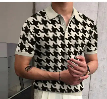 Person shown from shoulders to waist wearing a short-sleeve black-and-white houndstooth knit polo with a light collar and sleeve cuffs, standing indoors with hands clasped and a small forearm tattoo visible.