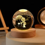 Glowing glass crystal ball lamp on a wooden base, etched with a small figure holding an umbrella and reaching toward a flower, with warm light and spark-like dots inside against a dark background.