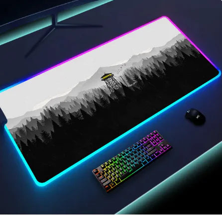 A gaming mouse pad with LED lighting along the edges, featuring a sunset landscape in orange and purple tones. A rainbow-colored mechanical keyboard and a gaming mouse are placed on the pad, creating a vibrant gaming setup.