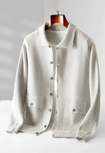 A white button-up knit cardigan with a classic collar, two front pockets with buttoned flaps, and ribbed cuffs and hem. The cardigan is layered over a white T-shirt and displayed on a wooden hanger, presenting a sophisticated and understated style.