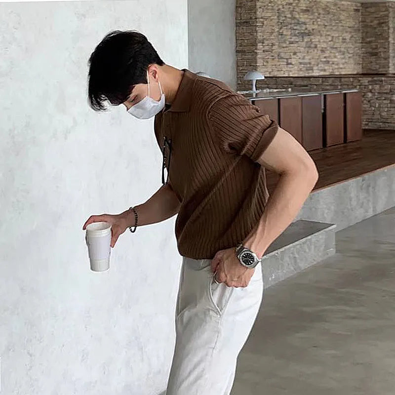 A fashionable man with a fit physique is captured in a candid moment, wearing a brown ribbed polo shirt with rolled-up sleeves, tucked into white trousers. He accessorizes with a silver wristwatch, a black bracelet, and a pair of glasses hanging from his shirt collar. He is also wearing a white face mask. Holding a white takeaway coffee cup in one hand, he places the other hand in his pocket while slightly bending forward. The background features a modern, minimalistic indoor space with a textured concrete