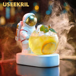 A clear glass cocktail drink glass with an astronaut figurine design on top, holding a glass of a beverage, with a blurred background suggesting a cosmic or space-themed setting.