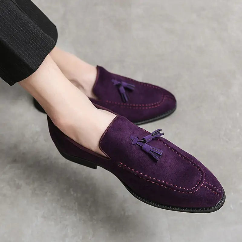 A pair of elegant burgundy leather loafers with a glossy finish. The shoes feature gold-toned metal horsebit detailing across the uppers and are displayed with wooden shoe trees inserted. They have a sleek, elongated silhouette with subtle stitching along the toe cap. The loafers are placed on a dark surface, partially framed by a wooden picture frame, emphasizing their classic and sophisticated style.