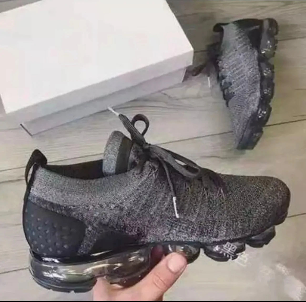 Hand holding a gray knit lace-up sneaker with a black heel and segmented air-cushion sole, with a white shoebox and the other shoe on a light wood floor in the background.