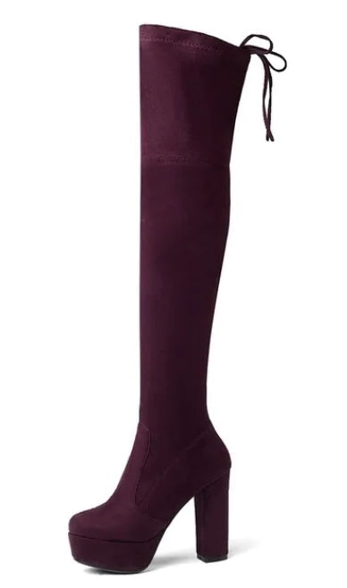 A woman wearing black high-heeled, over-the-knee boots with an open lattice cutout design running up the back, revealing skin between the horizontal straps. She stands indoors on a light wooden floor near a white ornate console table.