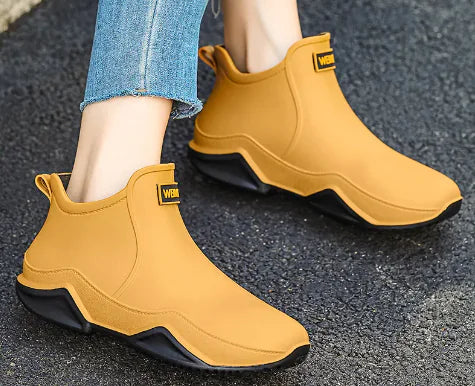 A pair of black waterproof ankle rain boots worn with cuffed jeans. The boots have a sleek, modern design with thick, textured soles and a branded label on the front that reads "WEMAM." The setting is an outdoor pavement, suggesting the boots are suited for wet or rainy weather.