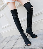 A close-up view of a person wearing black, ripped, over-the-knee denim boots with peep toes and high heels, paired with frayed white shorts, standing on a stone pavement near a white wall.