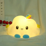 A glowing cloud-shaped night light with a cute face, blue feet, small yellow hands, and a rainbow accent on top, sitting on a bed near a book and red berries.