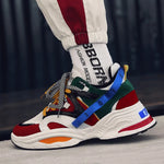 Close-up of a chunky multicolor sneaker (red, green, white, and blue) with patterned laces and a thick white sole, worn with white socks and track pants.