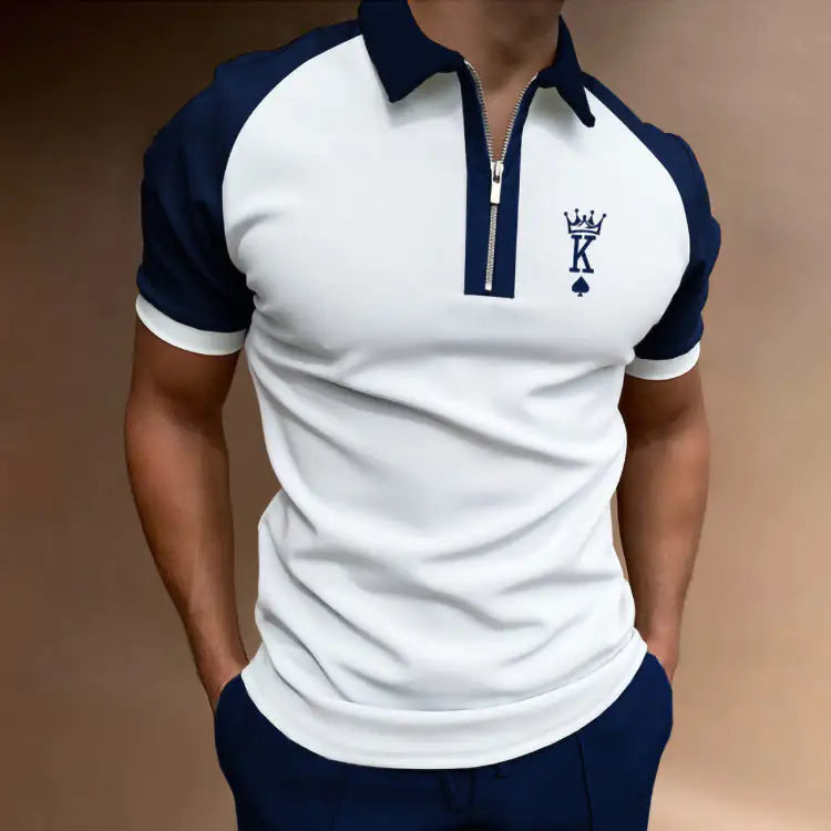 A man wearing a white polo shirt with navy sleeves and collar, a short zipper placket, and a small “K” crown logo on the chest, paired with navy pants against a plain background.