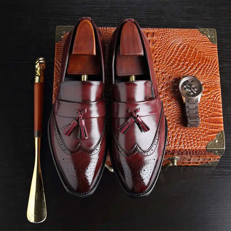 A pair of elegant burgundy leather loafers with a glossy finish. The shoes feature gold-toned metal horsebit detailing across the uppers and are displayed with wooden shoe trees inserted. They have a sleek, elongated silhouette with subtle stitching along the toe cap. The loafers are placed on a dark surface, partially framed by a wooden picture frame, emphasizing their classic and sophisticated style.