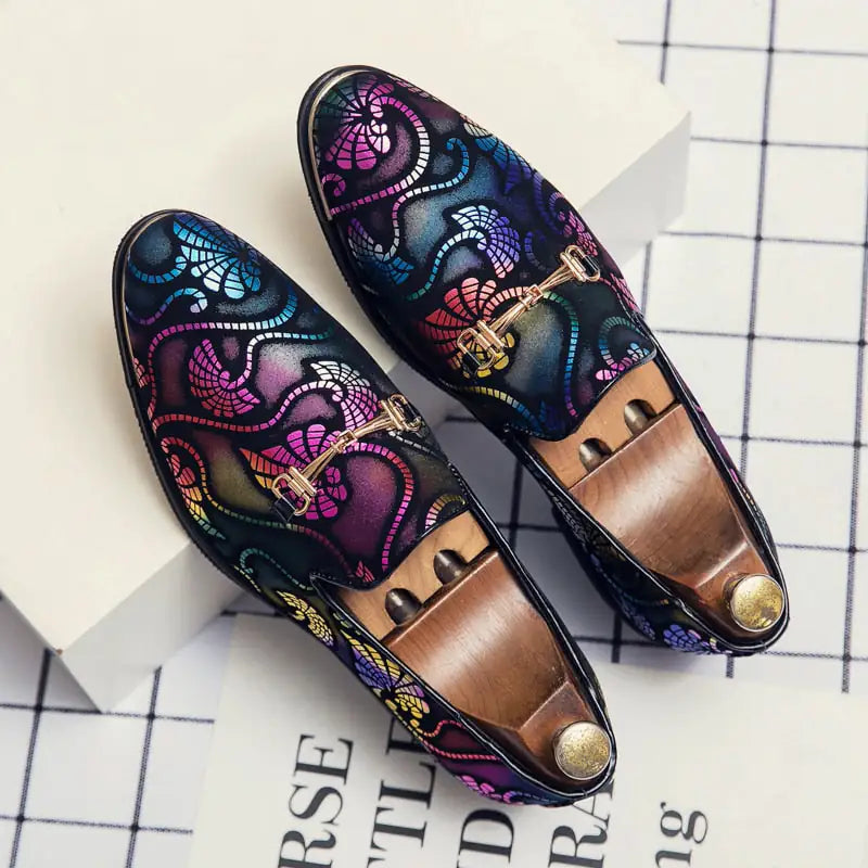 A pair of elegant burgundy leather loafers with a glossy finish. The shoes feature gold-toned metal horsebit detailing across the uppers and are displayed with wooden shoe trees inserted. They have a sleek, elongated silhouette with subtle stitching along the toe cap. The loafers are placed on a dark surface, partially framed by a wooden picture frame, emphasizing their classic and sophisticated style.