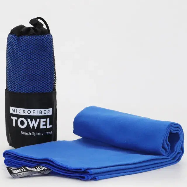 A microfiber towel rolled up next to its mesh carry bag. The towel is bright blue with a "Microfiber Towel" label on the bag, labeled for beach, sports, and travel use. The towel is unrolled slightly, showing its soft texture