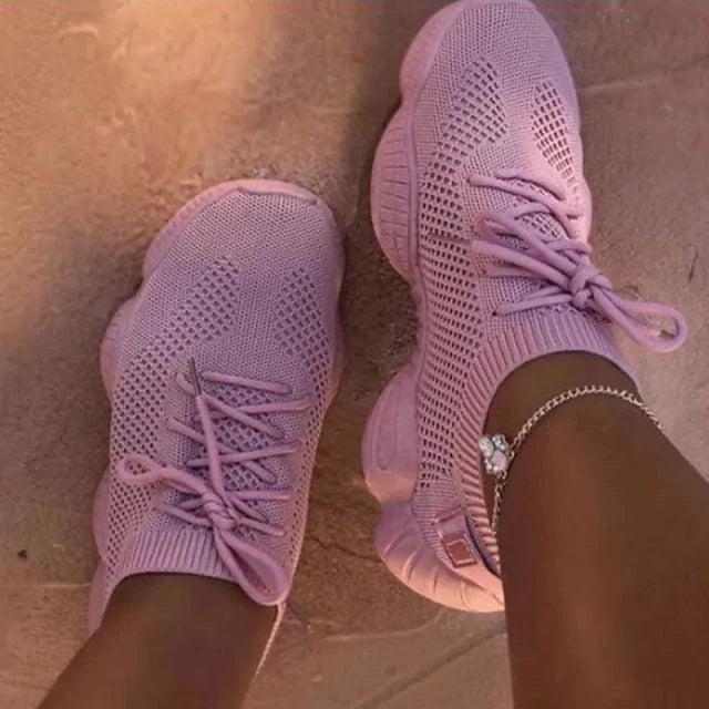 A pair of stylish white knitted athletic sneakers worn by a person standing on an asphalt surface. The shoes feature a breathable mesh design, thick cushioned soles, and gray laces. The person is also wearing white pants and a gold anklet.