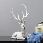 White decorative deer figurine with large branching antlers resting on a tabletop against a gray wall, next to a candle box and a folded blue cloth.
