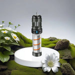 A compact cylindrical camping lantern with a black top and clear tube featuring an orange spiral, standing upright on a white display pedestal surrounded by green moss and white daisies.