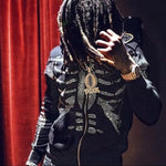 Person wearing a black zip-up hoodie with a rhinestone skeleton ribcage design, long dreadlocks covering their face, holding their hair, and wearing a large pendant chain and gold watch in front of a red curtain.