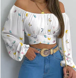 Woman wearing a white off-shoulder long-sleeve crop top with colorful butterfly print, paired with high-waisted blue jeans and a wide beige belt with a rhinestone buckle, standing against a plain wall.