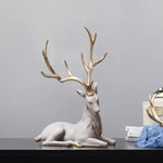 Decorative white reclining deer statue with large gold antlers, displayed on a dark tabletop against a plain gray wall, with a small stack of books and a blue cloth nearby.