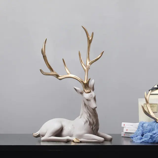 Decorative white reclining deer statue with large gold antlers, displayed on a dark tabletop against a plain gray wall, with a small stack of books and a blue cloth nearby.
