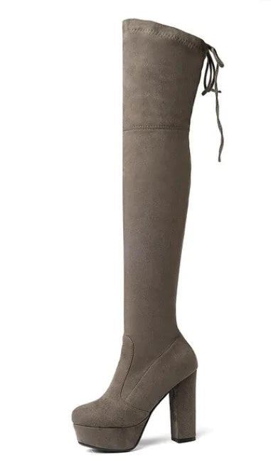 A woman wearing black high-heeled, over-the-knee boots with an open lattice cutout design running up the back, revealing skin between the horizontal straps. She stands indoors on a light wooden floor near a white ornate console table.