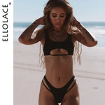 Woman standing on a beach wearing a black bikini with a scoop-neck top featuring a front cutout and high-cut bottoms with double side straps; ocean in the background and “ELLOLACE” text along the left edge.