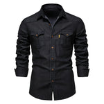A product image of a black long-sleeve button-up shirt (denim/workwear style) on a white background. It has a pointed collar, brown/gold buttons, and two chest flap pockets with button closures. The sleeves are shown slightly rolled/cuffed, and there’s a small vertical tag/label near the right chest pocket.
