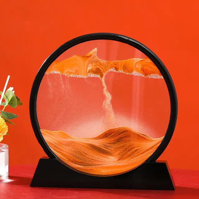 Circular desktop sand art frame on a black stand, showing orange sand forming layered dunes inside the glass against a bright orange background, with a small flower vase partly visible on the left.