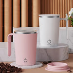 Two automatic self-stirring magnetic mugs, one pink and one white, with their respective mixing mechanisms visible. They are placed on a table with coffee beans scattered around and a flower vase in the background.