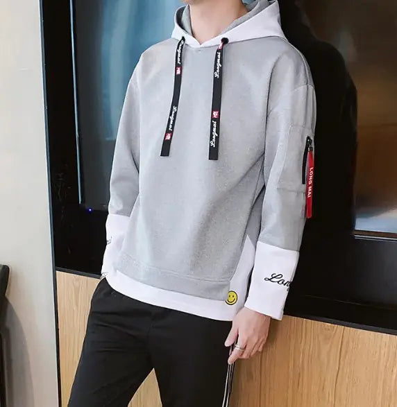 A stylish young man leans casually against a wooden wall while wearing a modern hoodie with black accents. The hoodie features black drawstrings with white text, black cuffs with embroidered details, and a red fabric tag on the left sleeve with 'LONG MA' printed on it. He is also wearing black athletic pants with white stripes on the sides. His accessories include silver rings on his fingers. The background consists of a contemporary indoor setting with a mix of wood and concrete elements.