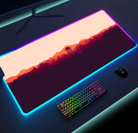 A gaming mouse pad with LED lighting along the edges, featuring a sunset landscape in orange and purple tones. A rainbow-colored mechanical keyboard and a gaming mouse are placed on the pad, creating a vibrant gaming setup.