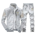 Light gray men’s tracksuit set with a zip-up jacket and matching jogger pants, featuring gold “AR” logos and ornate gold scroll patterns on the sleeves, shown on a white background.
