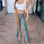 Woman wearing a white ribbed crop top paired with high-waisted, flared boho-style pants featuring a colorful vertical pattern in blue, orange, and yellow tones, posing barefoot indoors on a wooden floor.