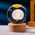 A clear glass/crystal sphere with an etched white dandelion design sits on a round wooden LED base, glowing warmly against a dark blue background with part of a wooden frame visible on the right.