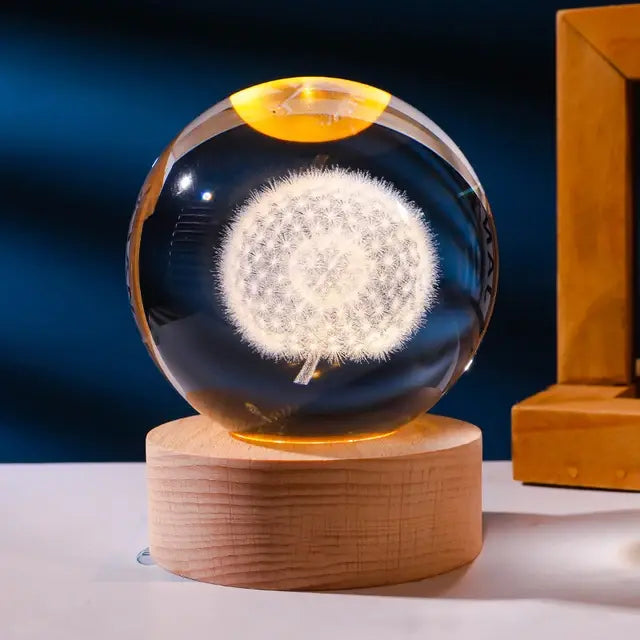 A clear glass/crystal sphere with an etched white dandelion design sits on a round wooden LED base, glowing warmly against a dark blue background with part of a wooden frame visible on the right.