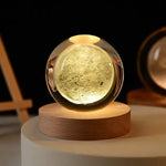 A glowing moon lamp in a clear glass orb, sitting on a wooden base, emitting a soft yellow light.