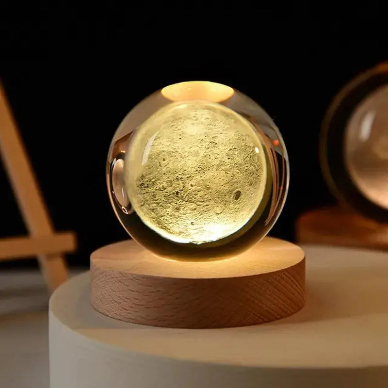 A glowing moon lamp in a clear glass orb, sitting on a wooden base, emitting a soft yellow light.