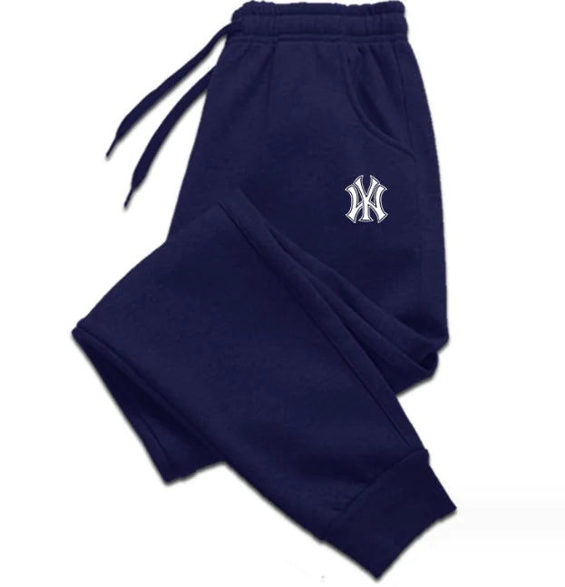 A pair of blue sweatpants with an elastic waistband and drawstrings, laid flat against a white background. The sweatpants feature ribbed cuffs at the ankles and side pockets. A black embroidered logo, resembling the New York Yankees emblem, is visible on the upper left thigh. The fabric appears to be soft, suggesting a comfortable and relaxed fit.