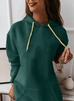 Woman wearing a long-sleeve, oversized maroon hoodie dress with a front kangaroo pocket and gold drawstrings. She is holding a light-colored handbag and posing indoors with a soft smile. The outfit has a casual, cozy look perfect for lounging or casual outings.