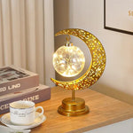 Gold crescent-moon table lamp with cutout patterns holding a glowing glass orb filled with fairy lights, sitting on a wooden desk beside books and a teacup.