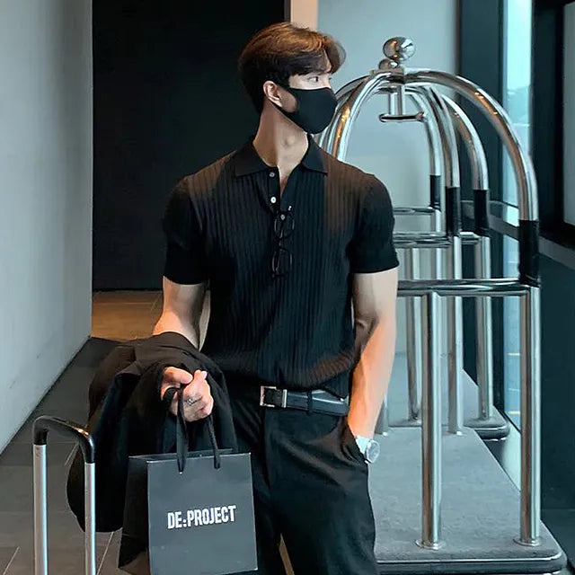 A stylish man with a fit physique is standing in a modern hallway near a luggage cart. He is dressed in an all-black outfit, consisting of a fitted ribbed polo shirt, trousers, and a black belt. He is wearing a black face mask and has sunglasses hanging from the neckline of his shirt. He carries a black shopping bag labeled 'DE: PROJECT' in one hand and a black jacket draped over his arm. His posture is relaxed, with one hand in his pocket, and he is looking to the side. The background features a sleek.