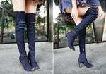 A close-up view of a person wearing black, ripped, over-the-knee denim boots with peep toes and high heels, paired with frayed white shorts, standing on a stone pavement near a white wall.